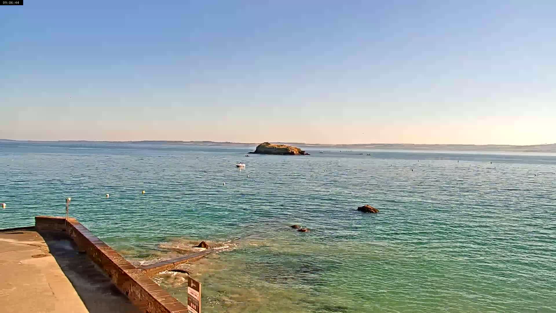 A clear, sunny day reveals a calm, teal ocean with a small island in the distance, viewed from a stone walkway leading to the water's edge.