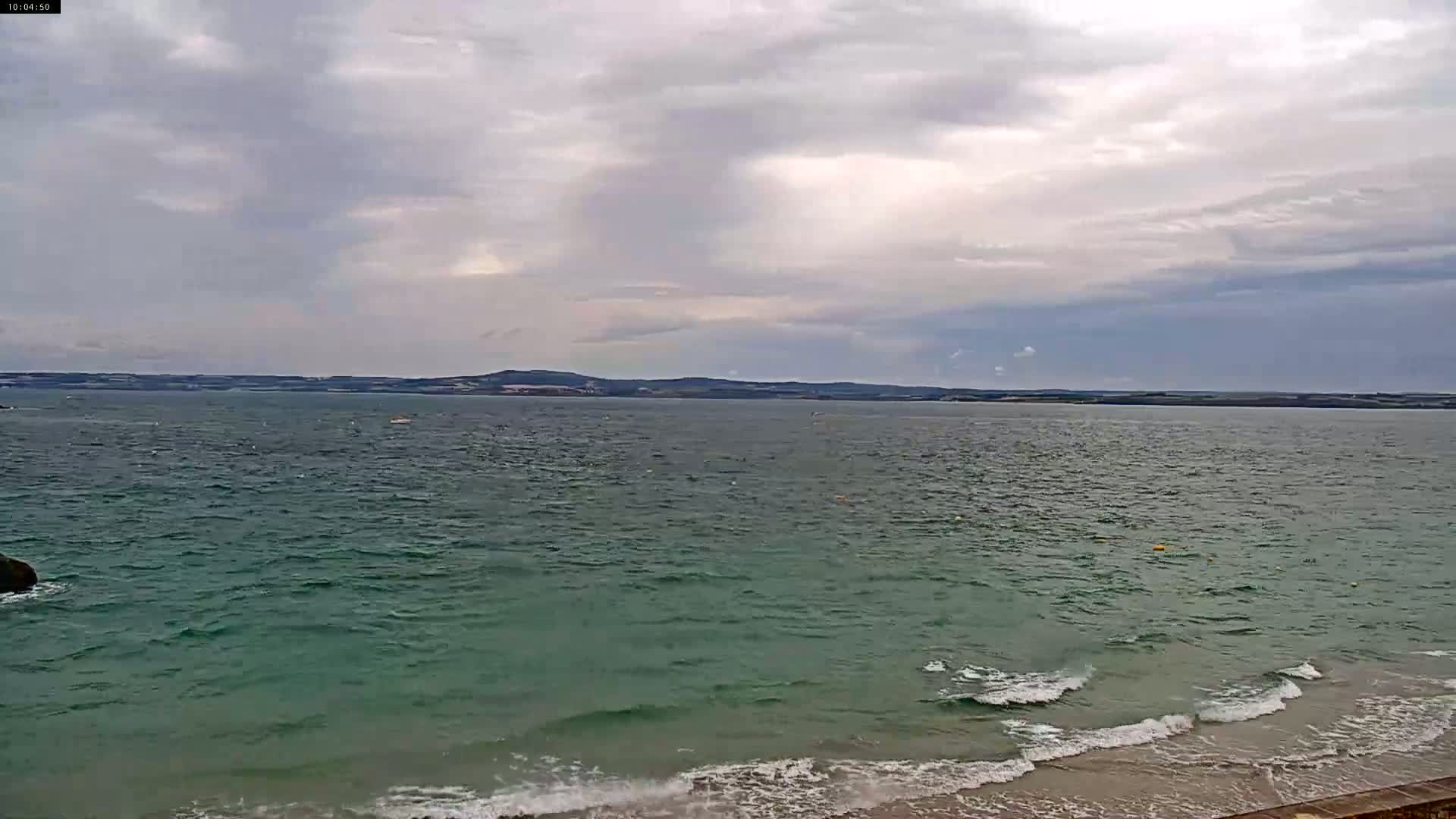A wide shot of a calm sea with small waves lapping a sandy shore under a cloudy sky.