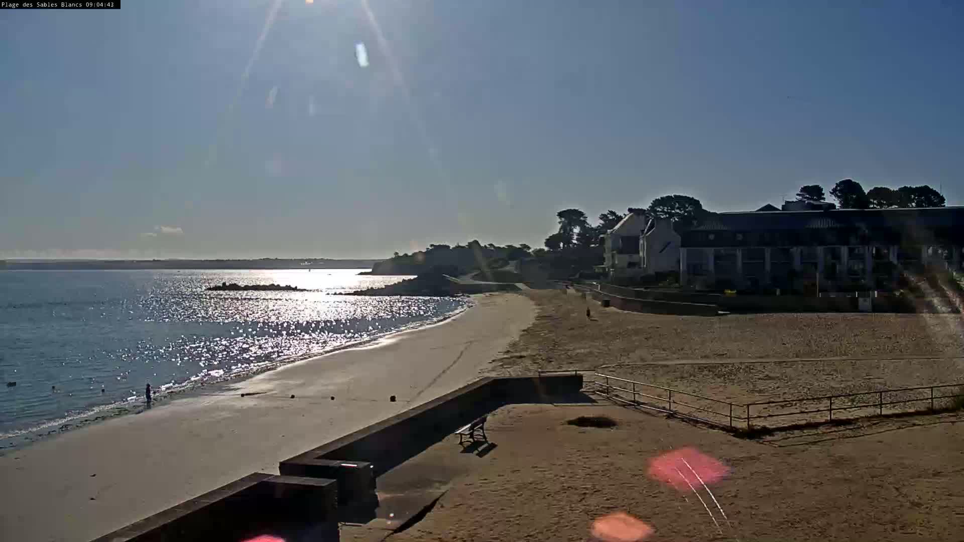 A sandy beach stretches out to a calm, sunlit sea under a clear blue sky, with a few buildings visible in the background.