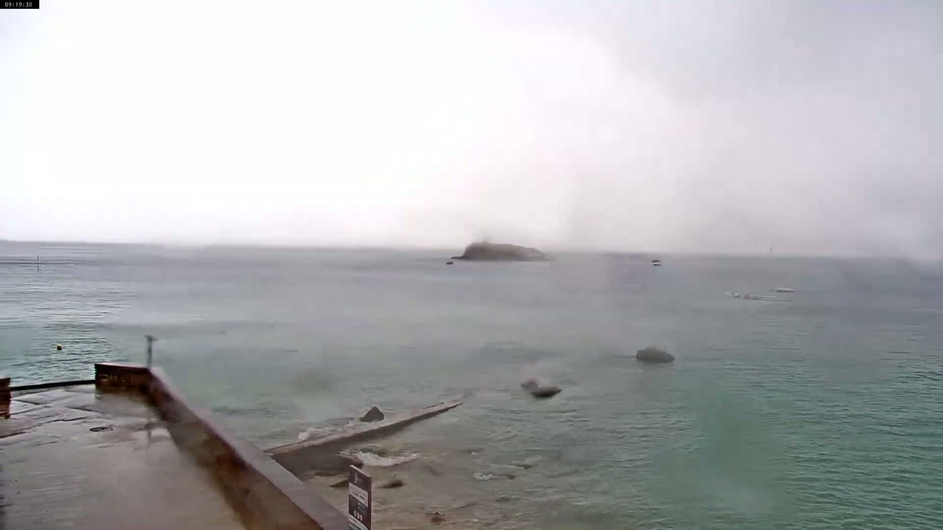 A wet, overcast day shows a rocky shoreline with a small island visible in the distance across a calm sea.