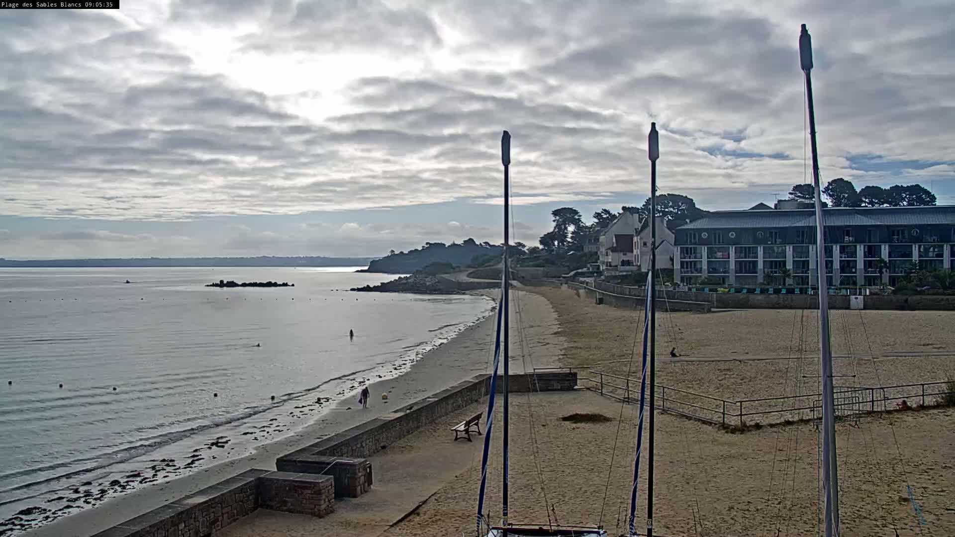 A sandy beach with calm water, a few people, and a multi-story building is visible under a mostly cloudy sky.