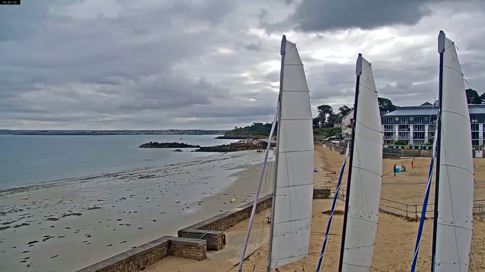 Three sailboats are on a sandy beach next to calm water under a cloudy sky.