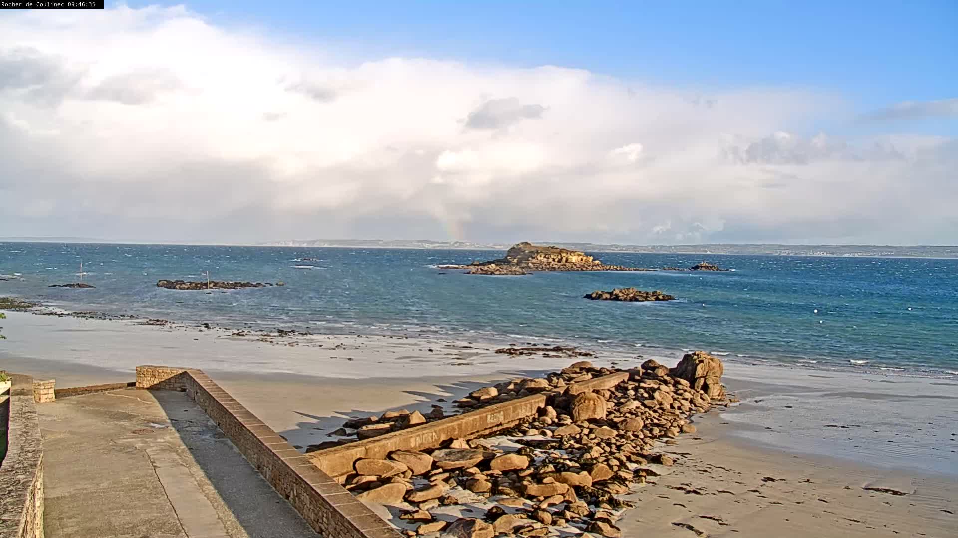 A bright, partially cloudy day with a faint rainbow over the horizon illuminates a vast blue-green ocean dotted with rocky islets, meeting a sandy beach with a rugged rocky shoreline and a stone-walled path in the foreground.