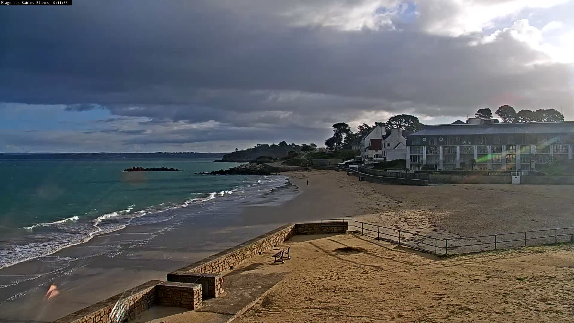 A wide sandy beach with turquoise waves crashing along the shore is backed by coastal buildings and a rocky headland under a sky of heavy, dark clouds with a contrasting burst of sunlight breaking through on the right.