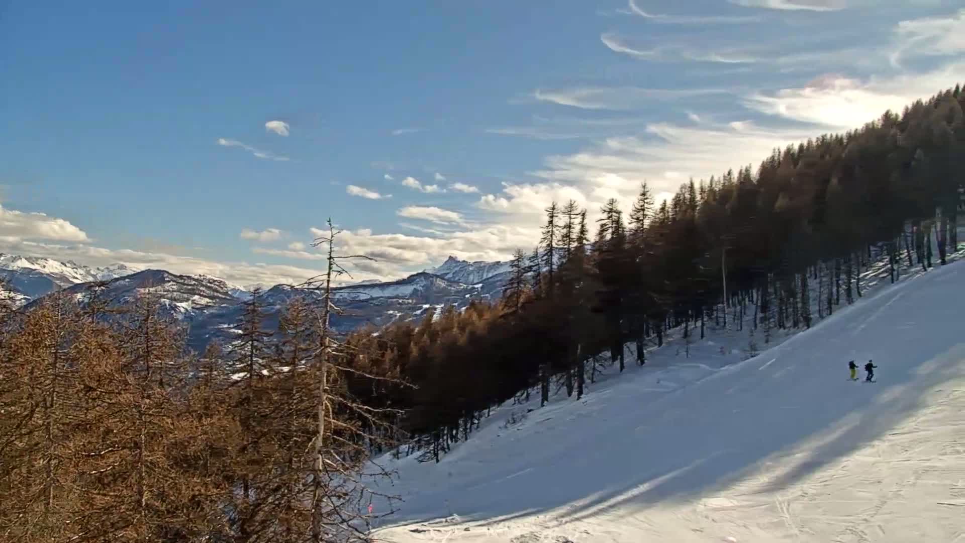 A snowy ski slope descends from a line of dark pine trees toward a distant, snow-capped mountain range under a mostly clear, bright blue sky with thin, wispy clouds.