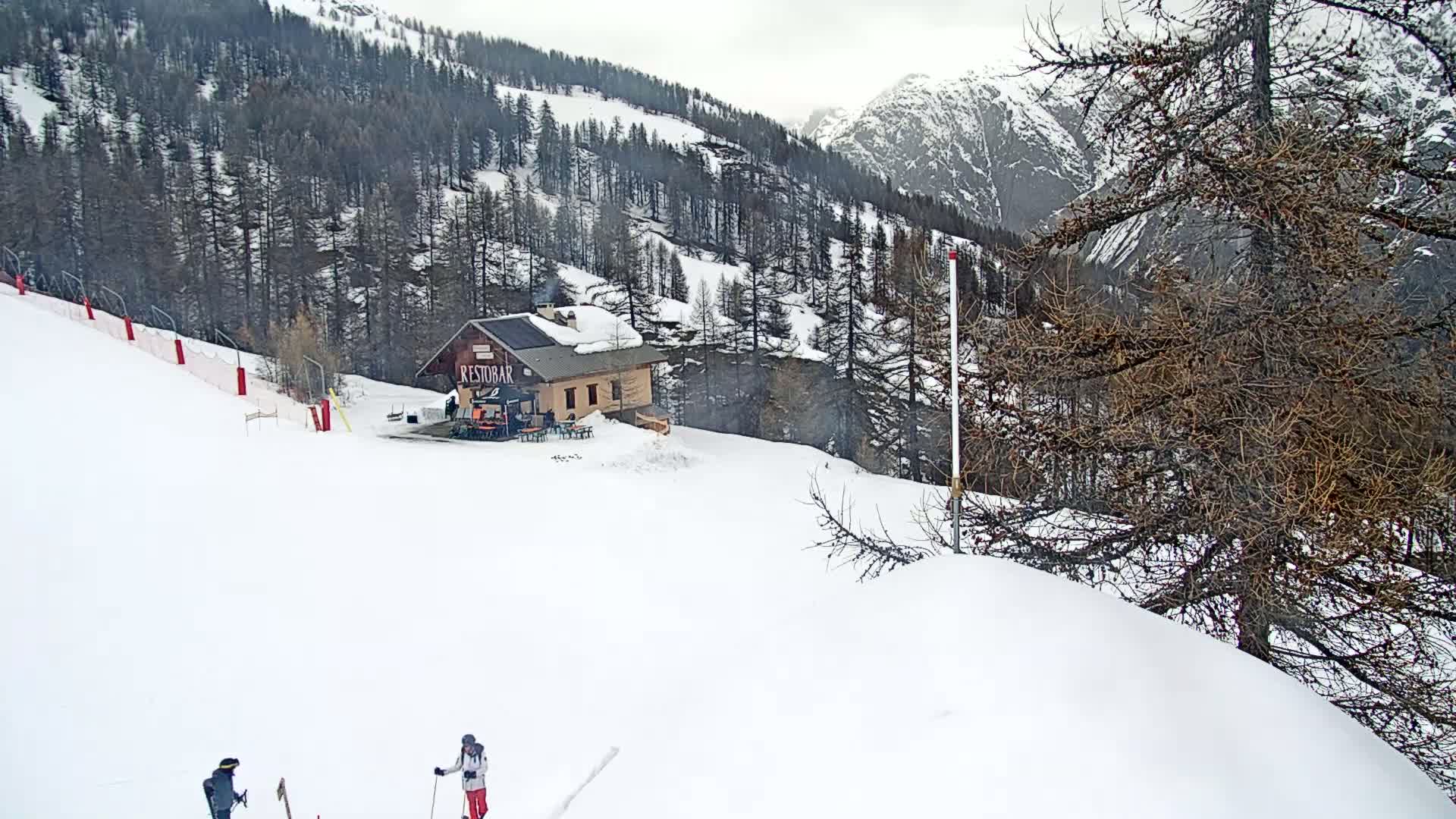 A snow-covered ski slope with a small building and two skiers, situated against a backdrop of a mountain range under an overcast sky.