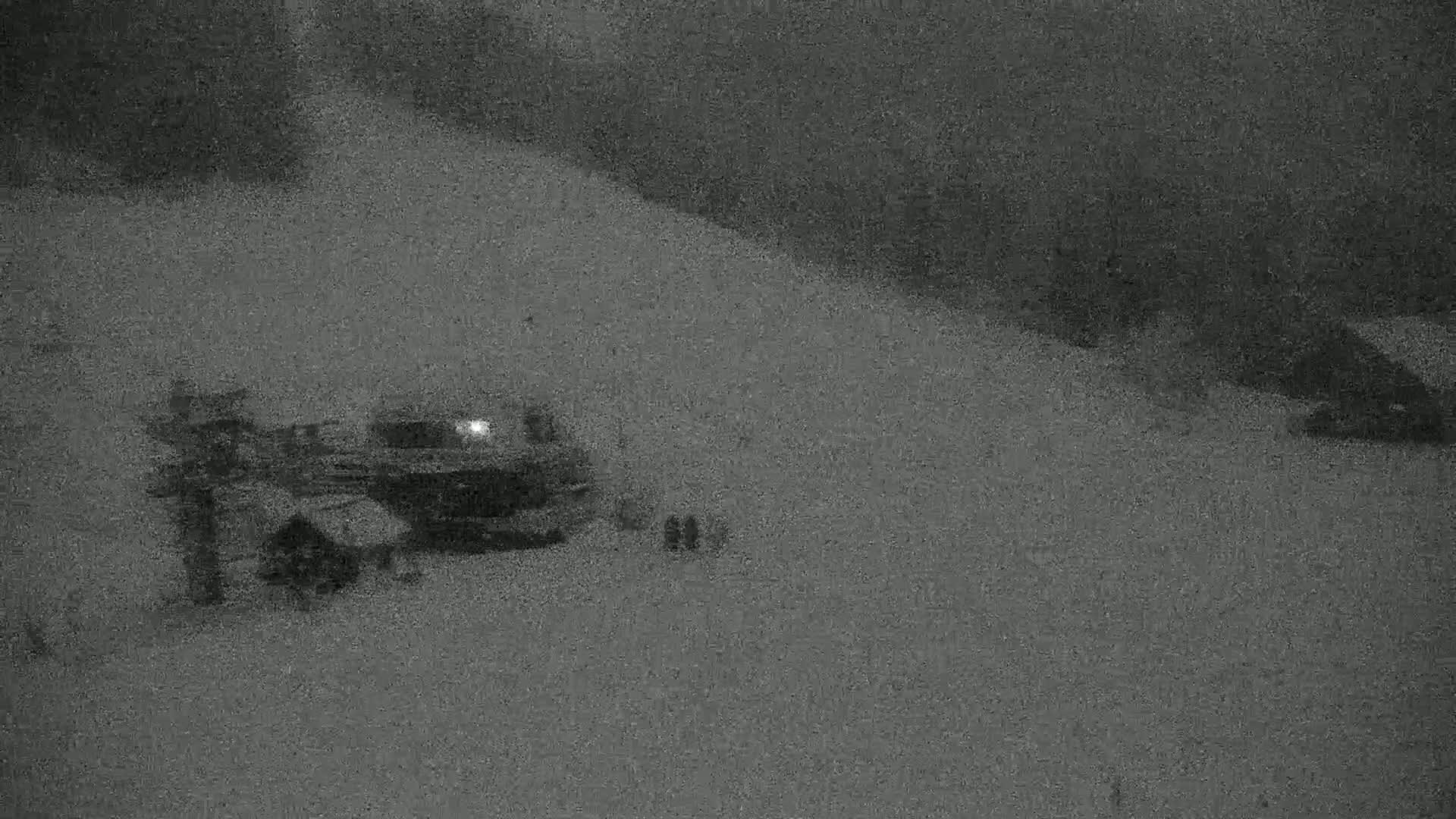 A snow-covered area at night shows a vehicle with its headlights on near some structures.
