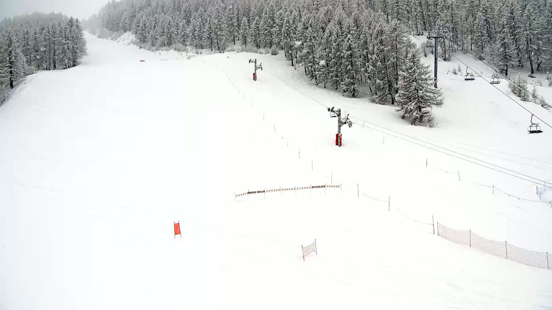 A snow-covered ski slope with several chairlifts and snow-covered trees in the background during a light snowfall.