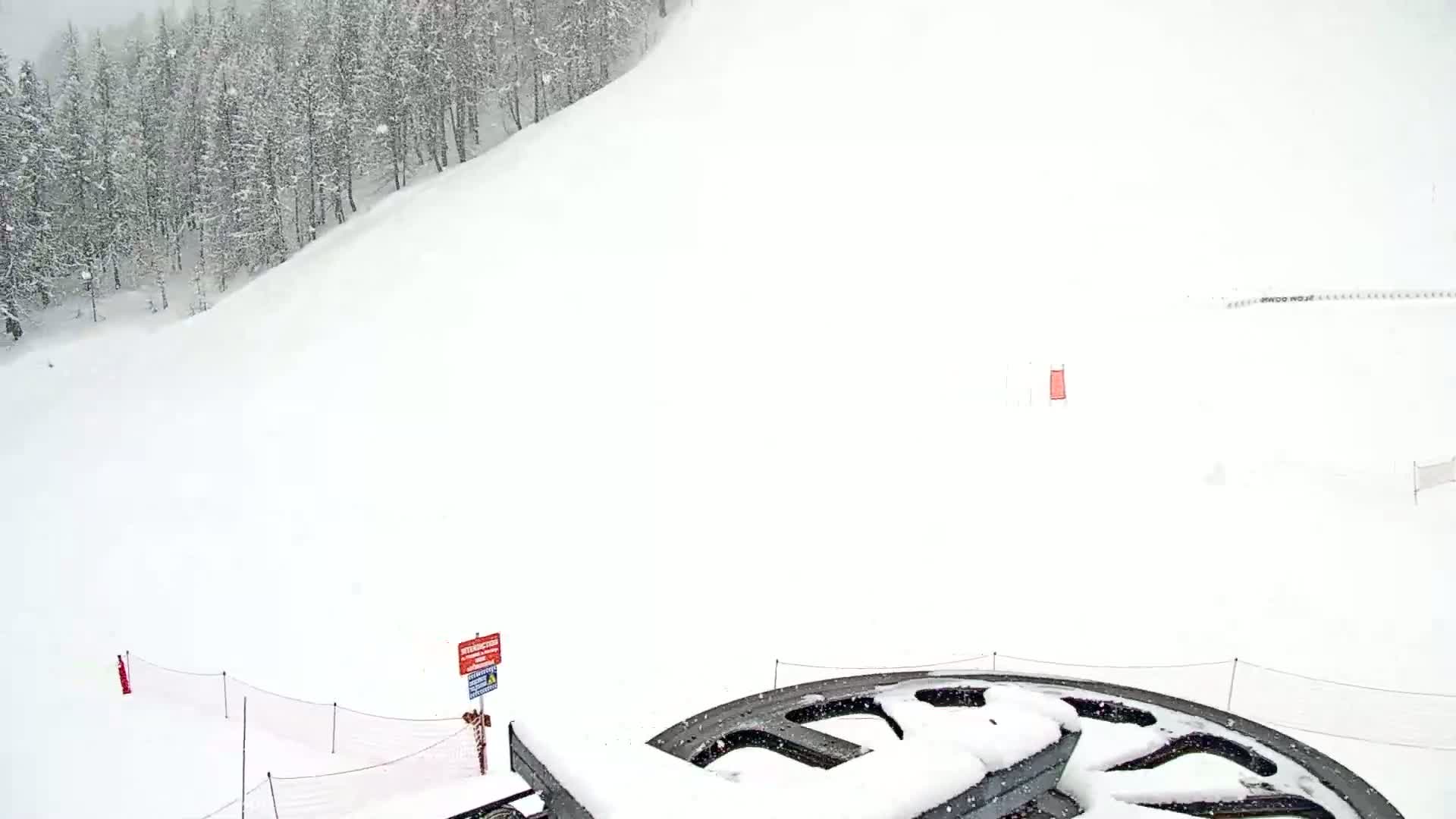 A snow-covered ski slope with a snow-covered ski lift pulley in the foreground, during a snowfall.