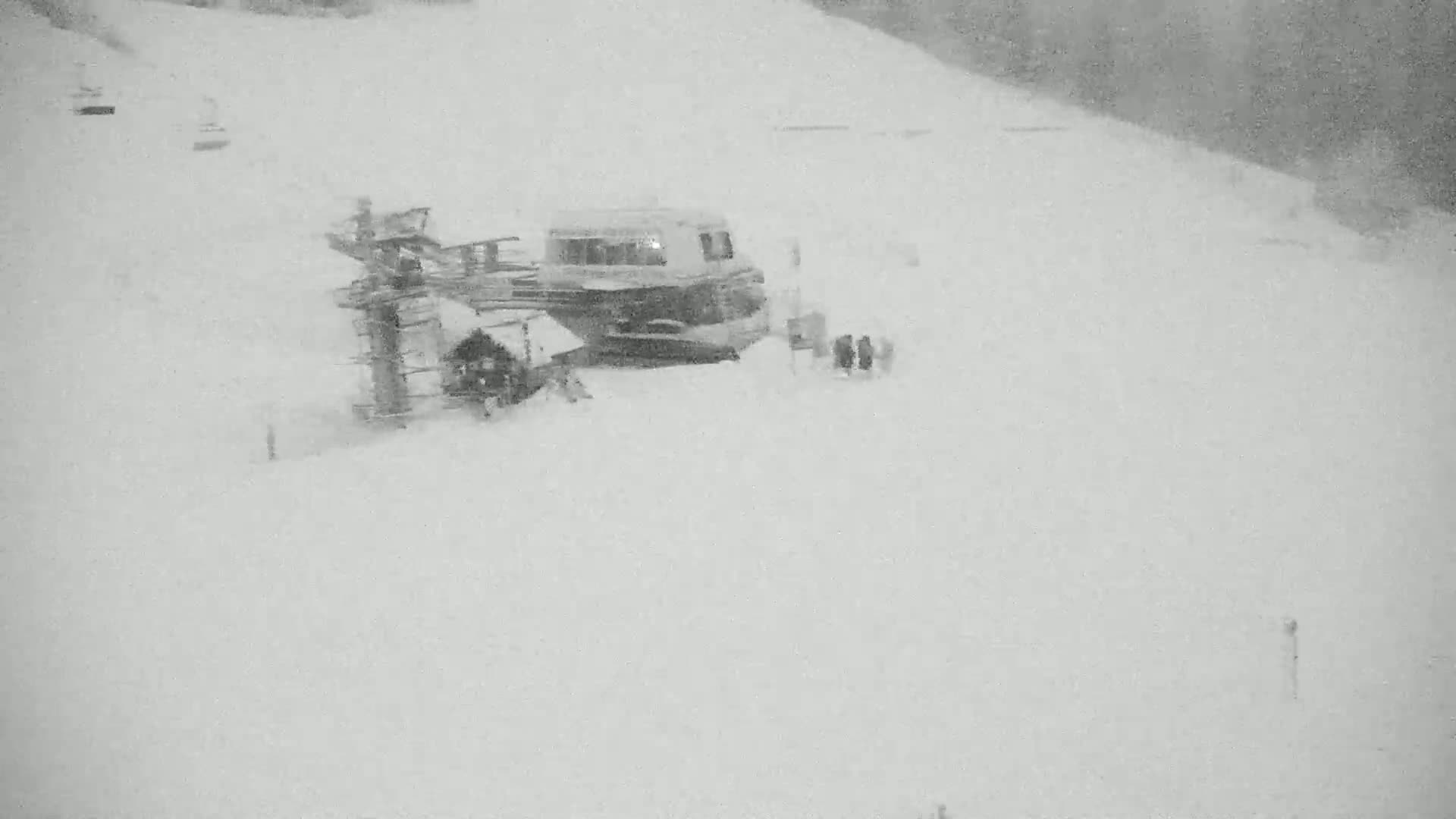 A snow-covered ski slope shows a ski lift, a small building, and a few people amidst a heavy snowfall.