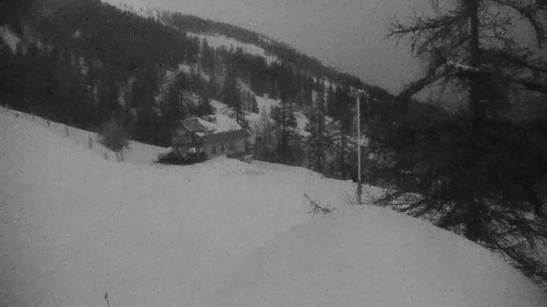 A small building sits on a snow-covered hillside, surrounded by dark evergreen trees under a gray, overcast sky.