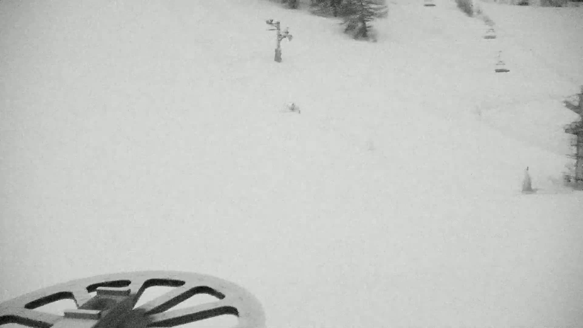 A snowy ski slope is viewed from a chairlift, showing a sparsely populated run and a ski lift in the distance.