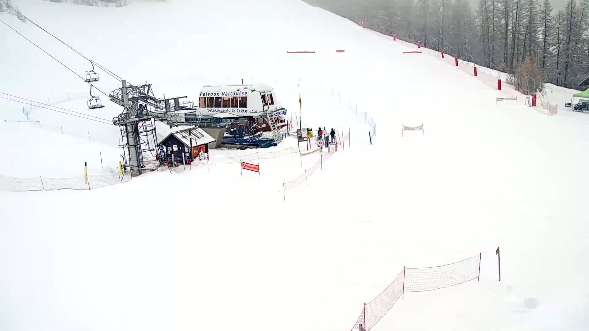 A ski lift and a small building are situated on a snow-covered ski slope during a light snowfall.