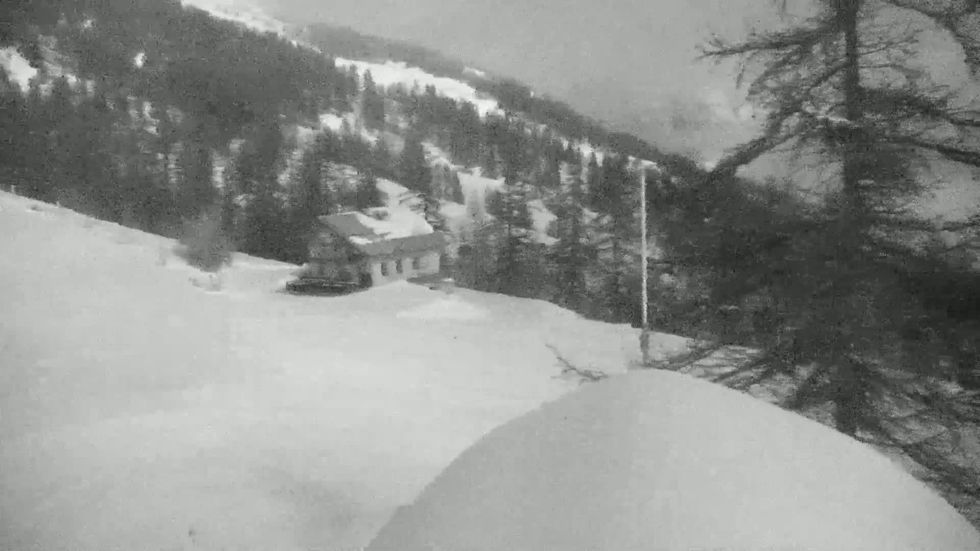 A snow-covered landscape featuring a small building nestled among evergreens on a hillside, with a snowy foreground and overcast weather conditions.