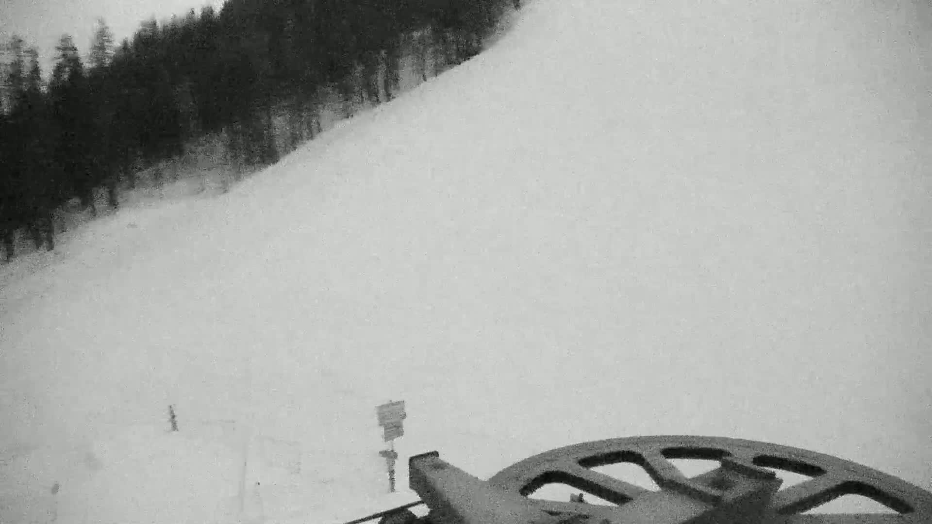 A snow-covered ski slope is viewed from a chairlift, with a dark forest visible in the background under an overcast sky.