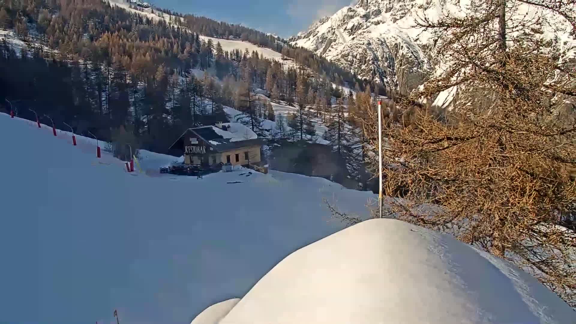 A snow-covered ski slope with a small building and trees, under a sunny sky with a mountainous backdrop.
