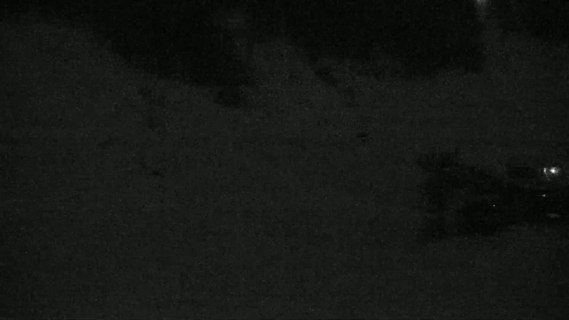 A low-light nighttime image shows a dark, grainy surface, possibly asphalt, with indistinct shapes and faint lights in the distance.