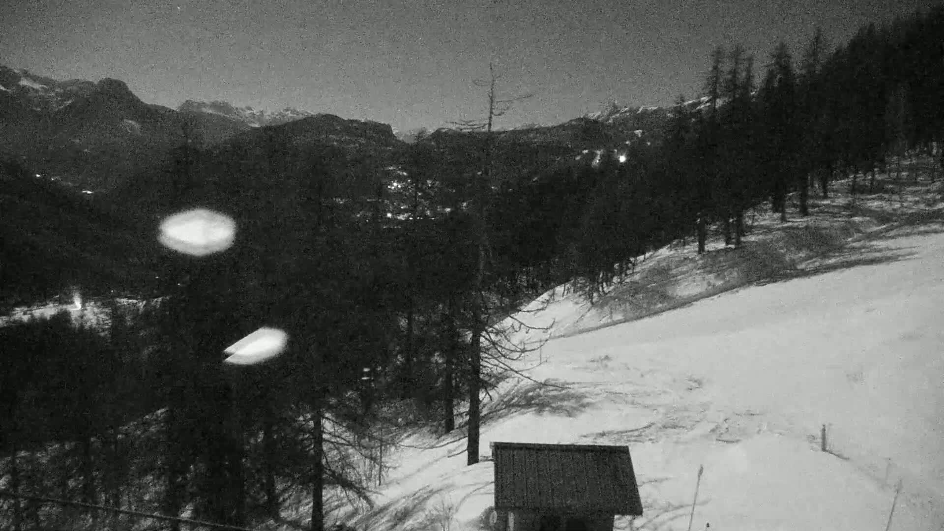A nighttime, snowy mountain landscape is seen from a high vantage point, showing a small structure in the foreground and a valley with distant lights beyond a dark forest.