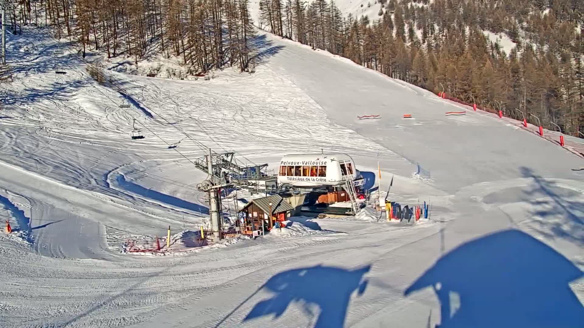 A ski lift station sits at the bottom of a snow-covered ski slope on a sunny day, surrounded by a coniferous forest.