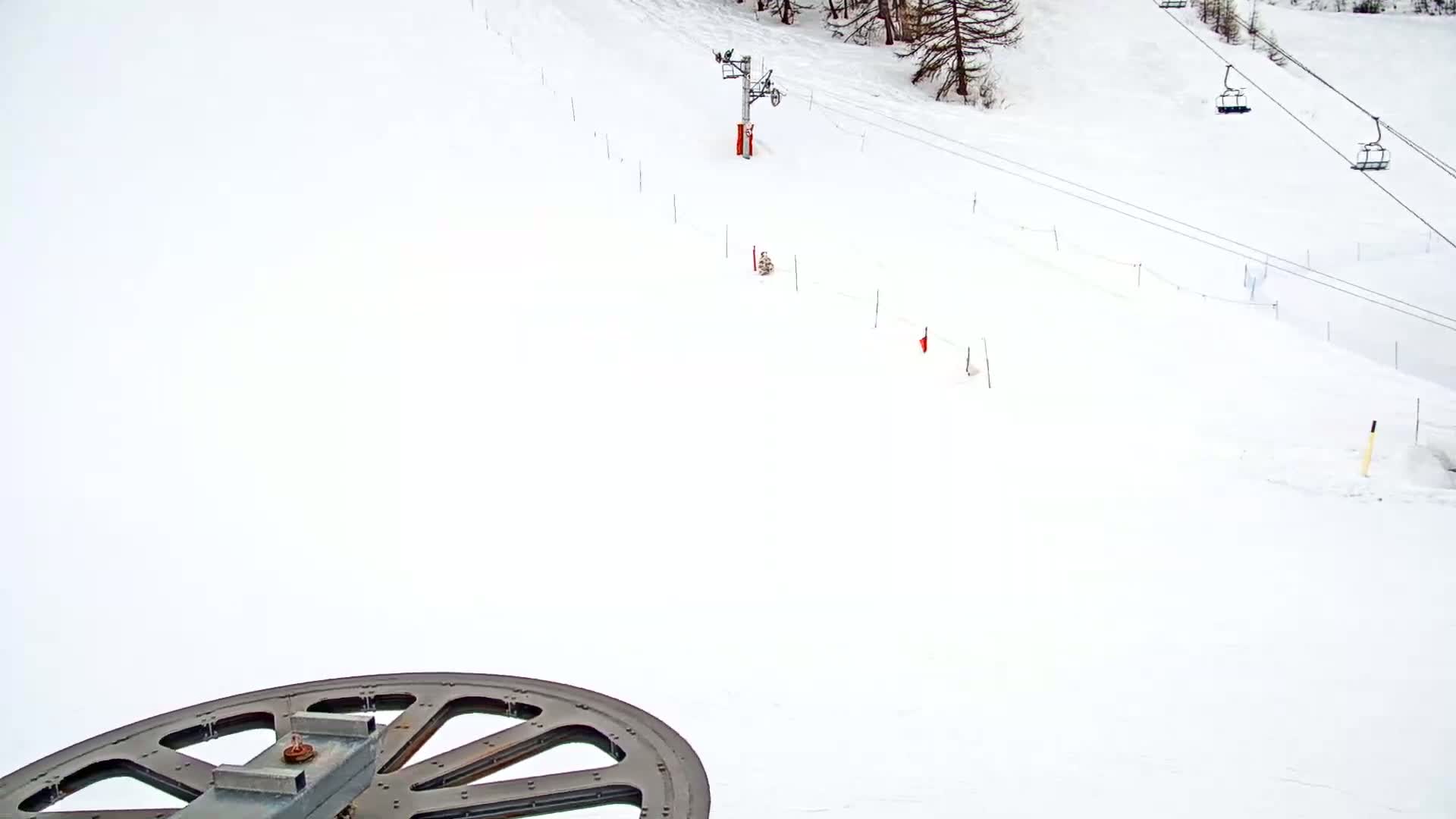 A large wheel of a ski lift is in the foreground of a snow-covered ski slope with two chairlifts and sparsely spaced markers.