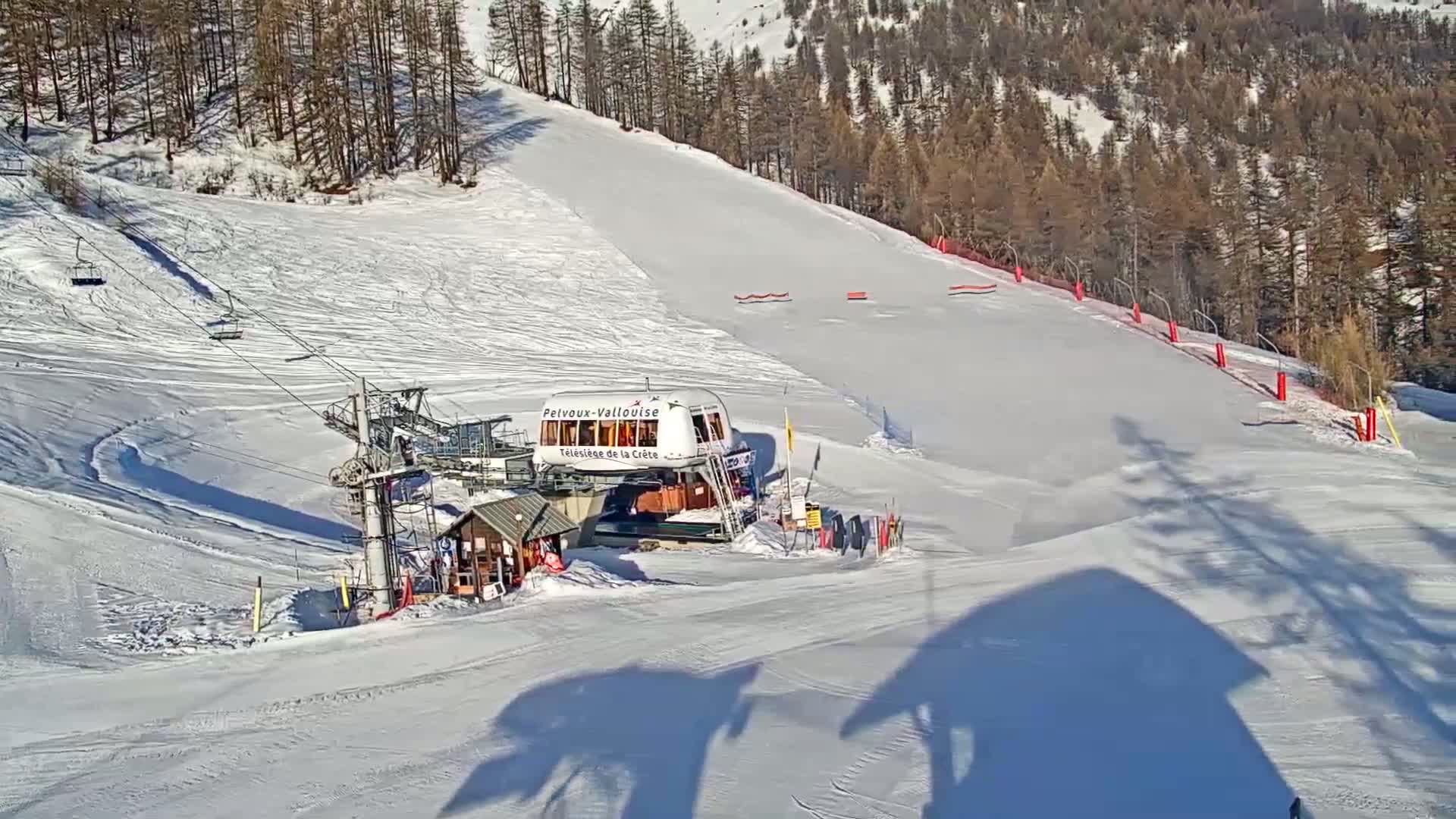 A ski lift station sits at the bottom of a snow-covered ski slope on a sunny day, with a forest of dark-green trees in the background.
