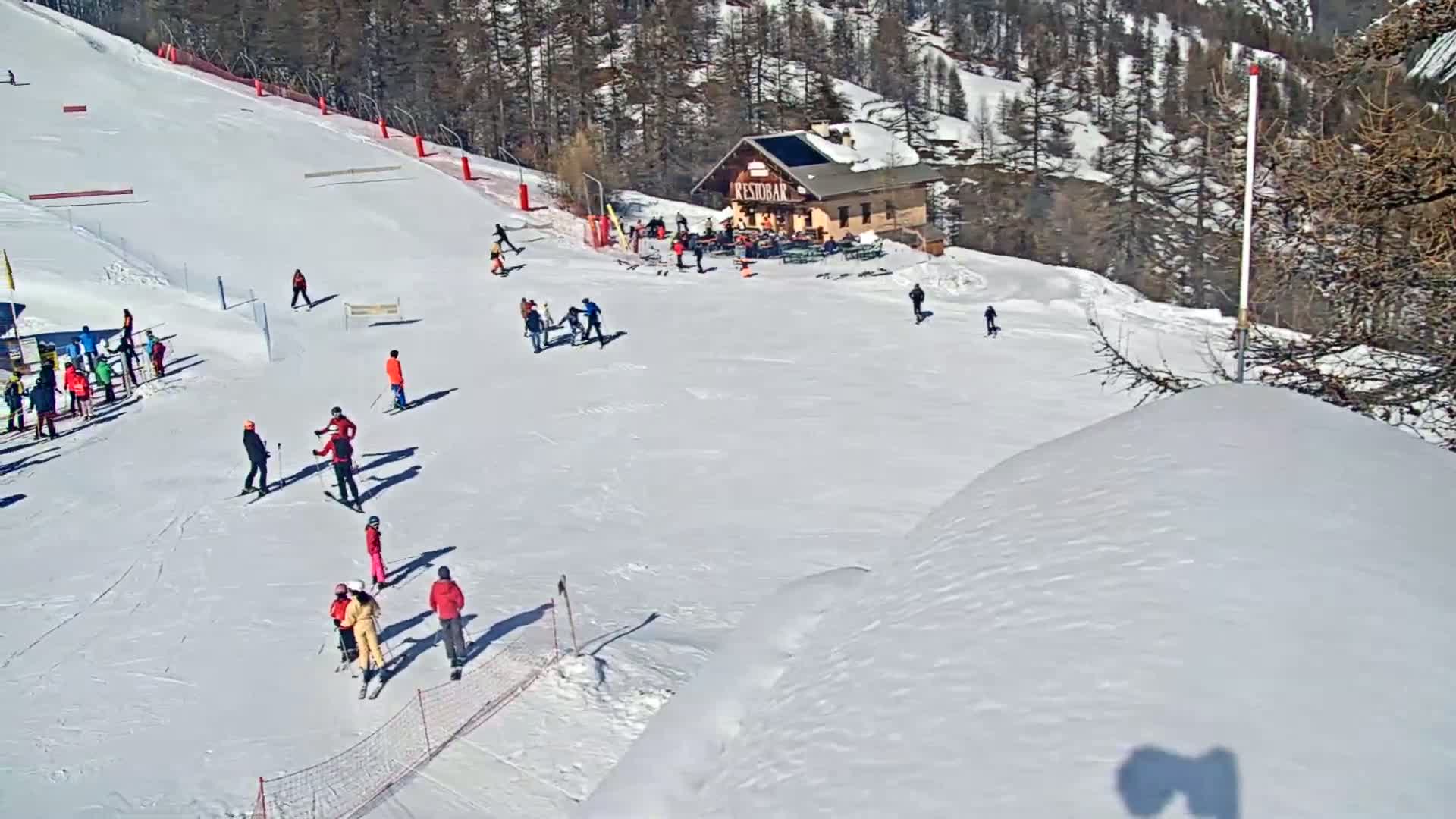 A ski slope with numerous skiers and snowboarders, a small lodge, and snowy, sunny conditions.