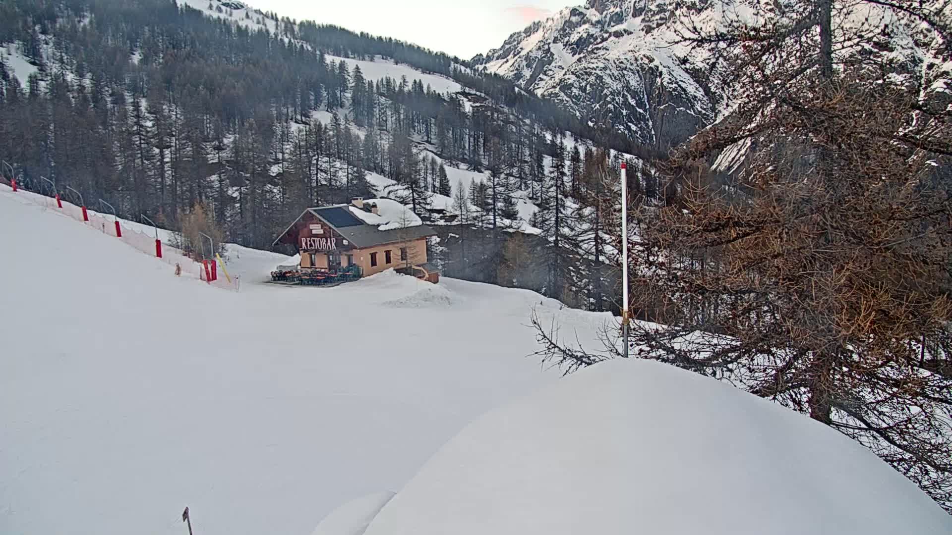 A snow-covered ski slope with a small building and trees in a mountainous area.
