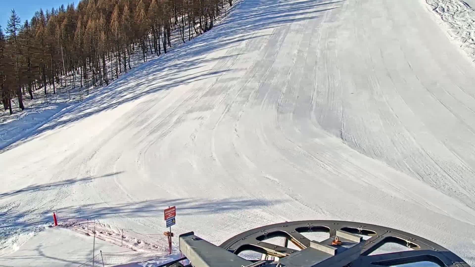 A snowy ski slope on a sunny day is seen from a ski lift, with a forest of dark trees in the background.