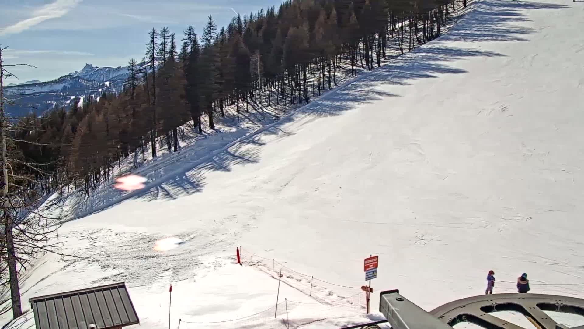 A snowy ski slope descends from a line of dark evergreen trees under a bright, sunny sky, with two people standing near the bottom.