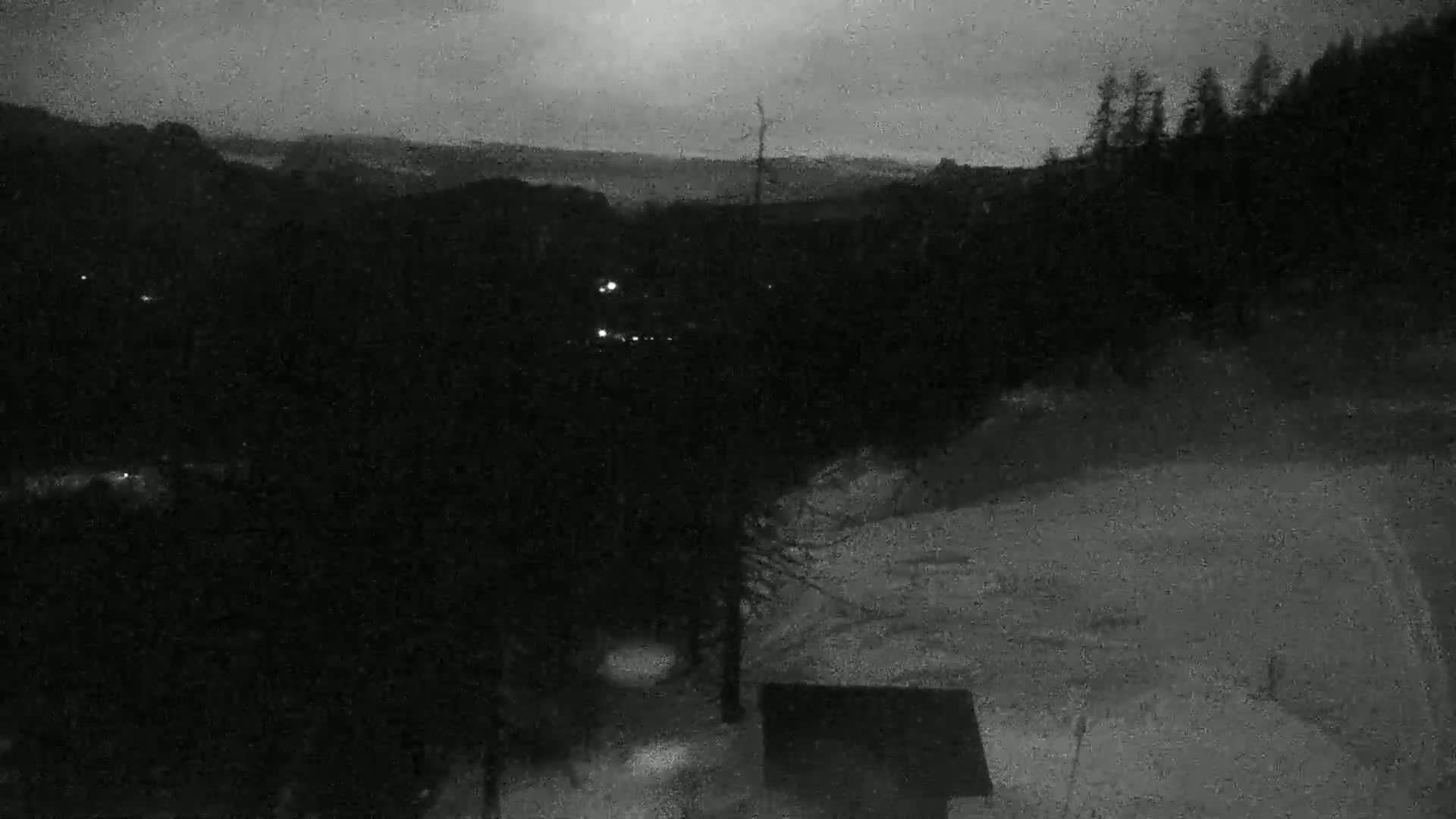 A dark, snowy landscape at night is visible, showing a hillside with a few distant lights and a small structure in the foreground under an overcast sky.