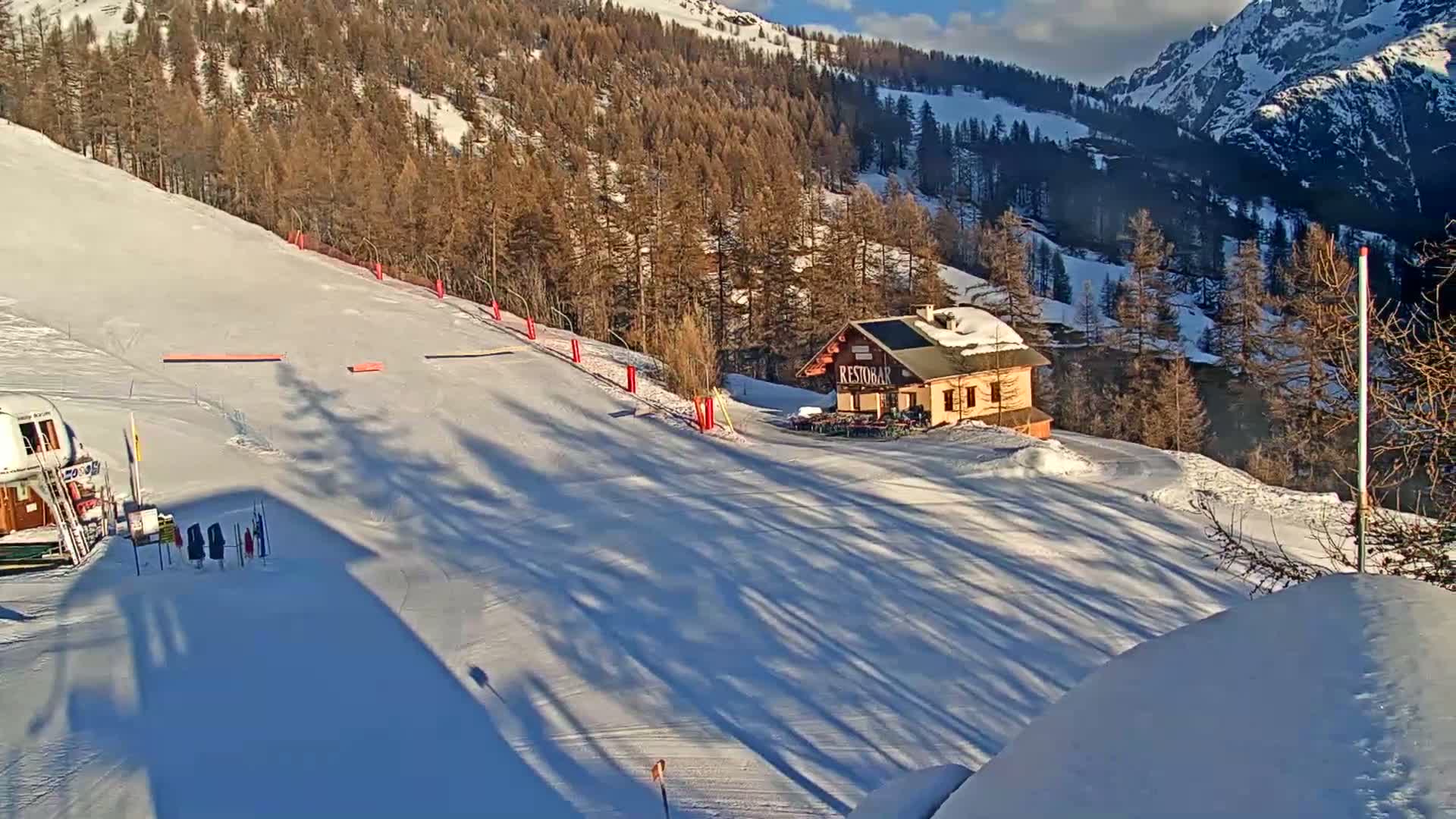 A snow-covered ski slope with a small building and surrounding pine trees on a sunny day.