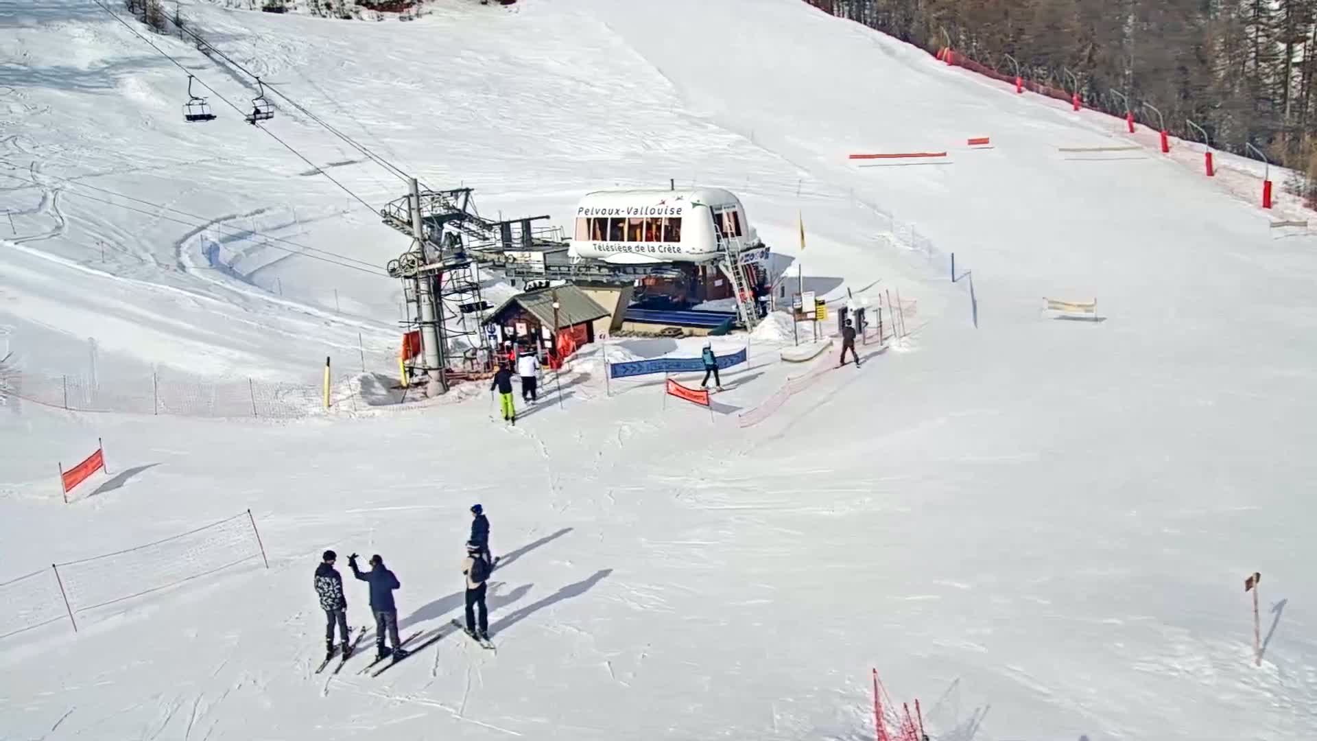 A ski lift station and several skiers are on a snowy slope on a sunny day.