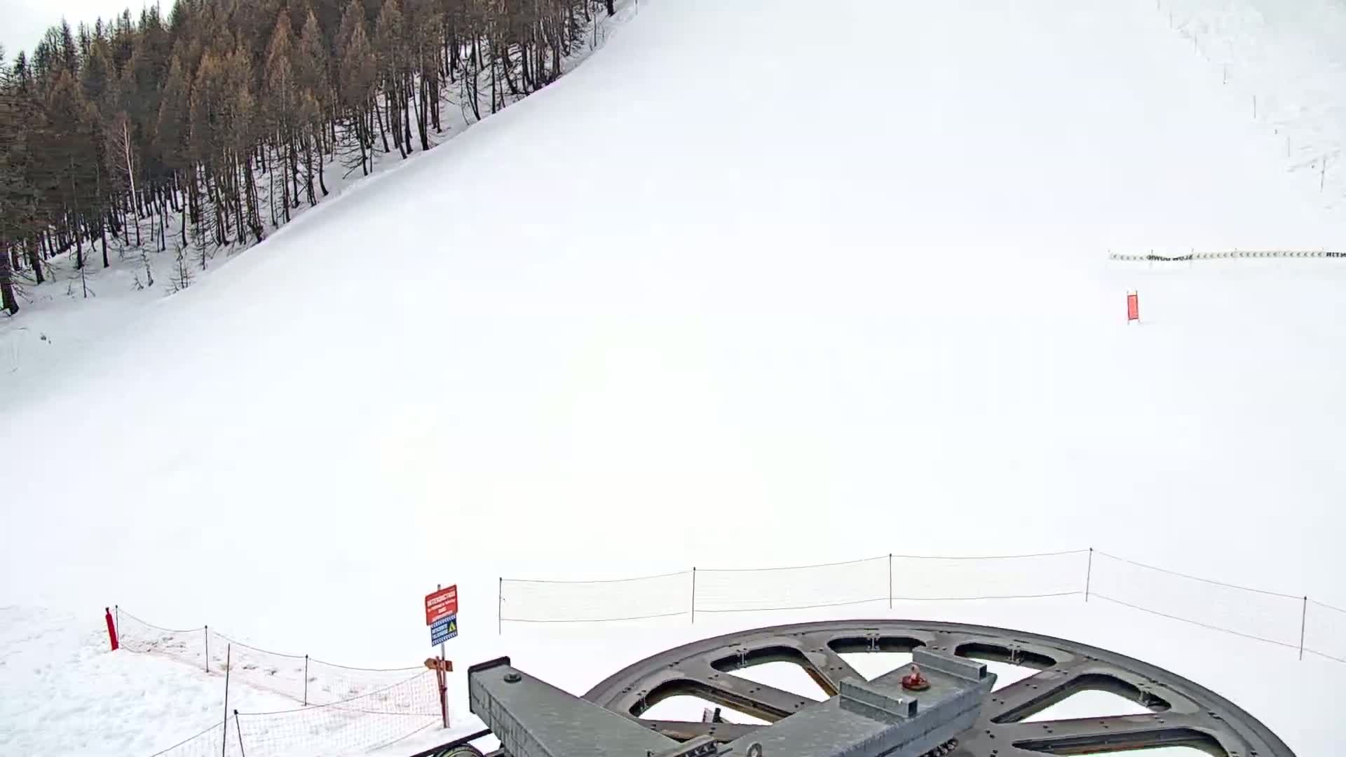 A large metal wheel is in the foreground of a snowy ski slope, with a dark forest visible in the background under a cloudy sky.