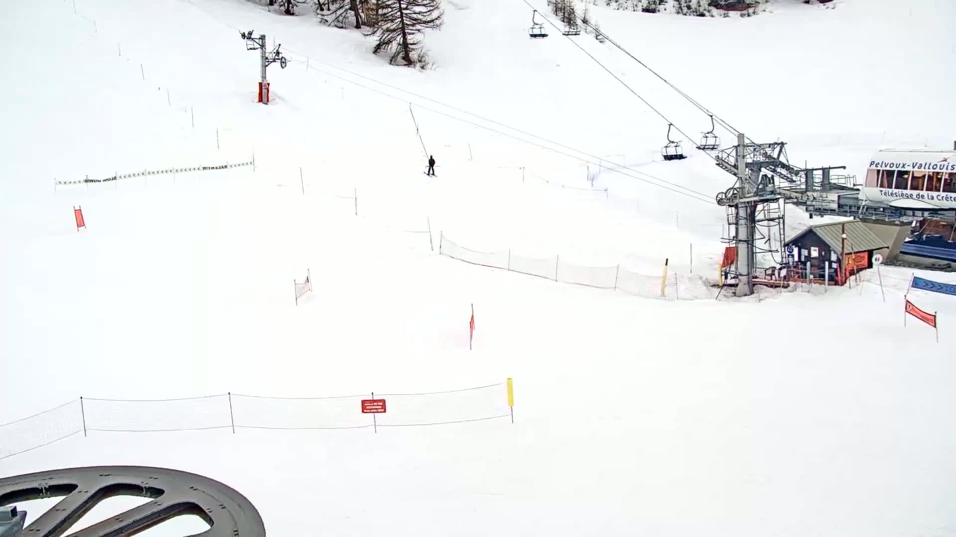 A snow-covered ski slope with a chairlift, a lone skier, and various safety barriers under a cloudy sky.