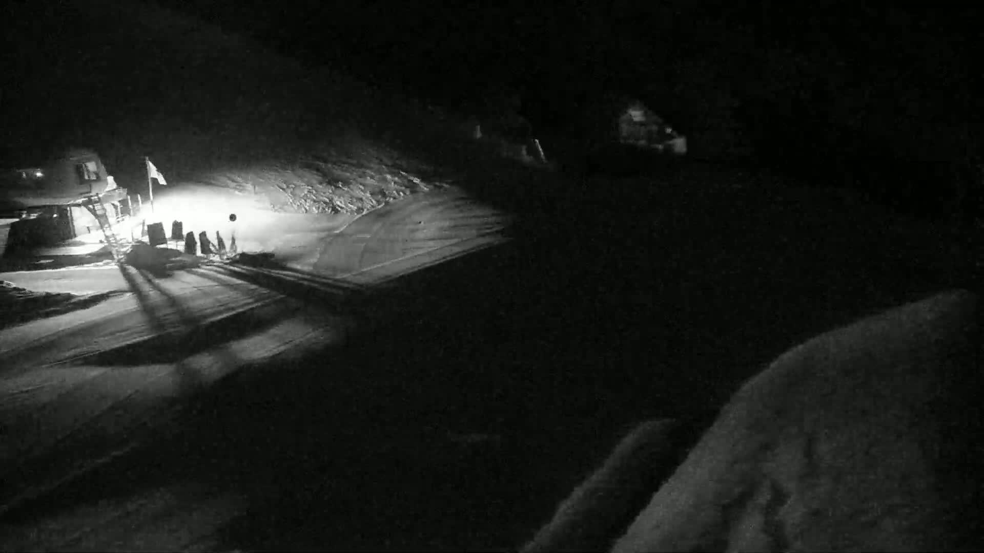 A nighttime, snow-covered scene shows a small building illuminated by a light, with long shadows cast across a flat snowy area.