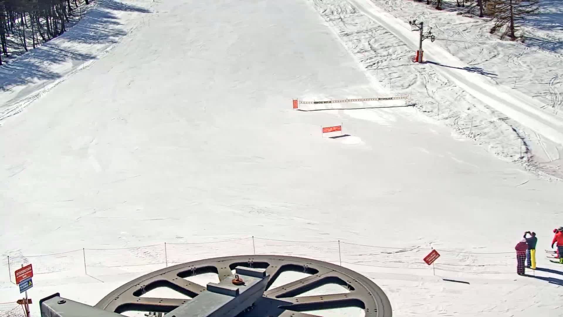 A snowy ski slope is viewed from above, showing a large metal wheel in the foreground, a few people standing near the bottom, and a ski lift in the background under sunny conditions.