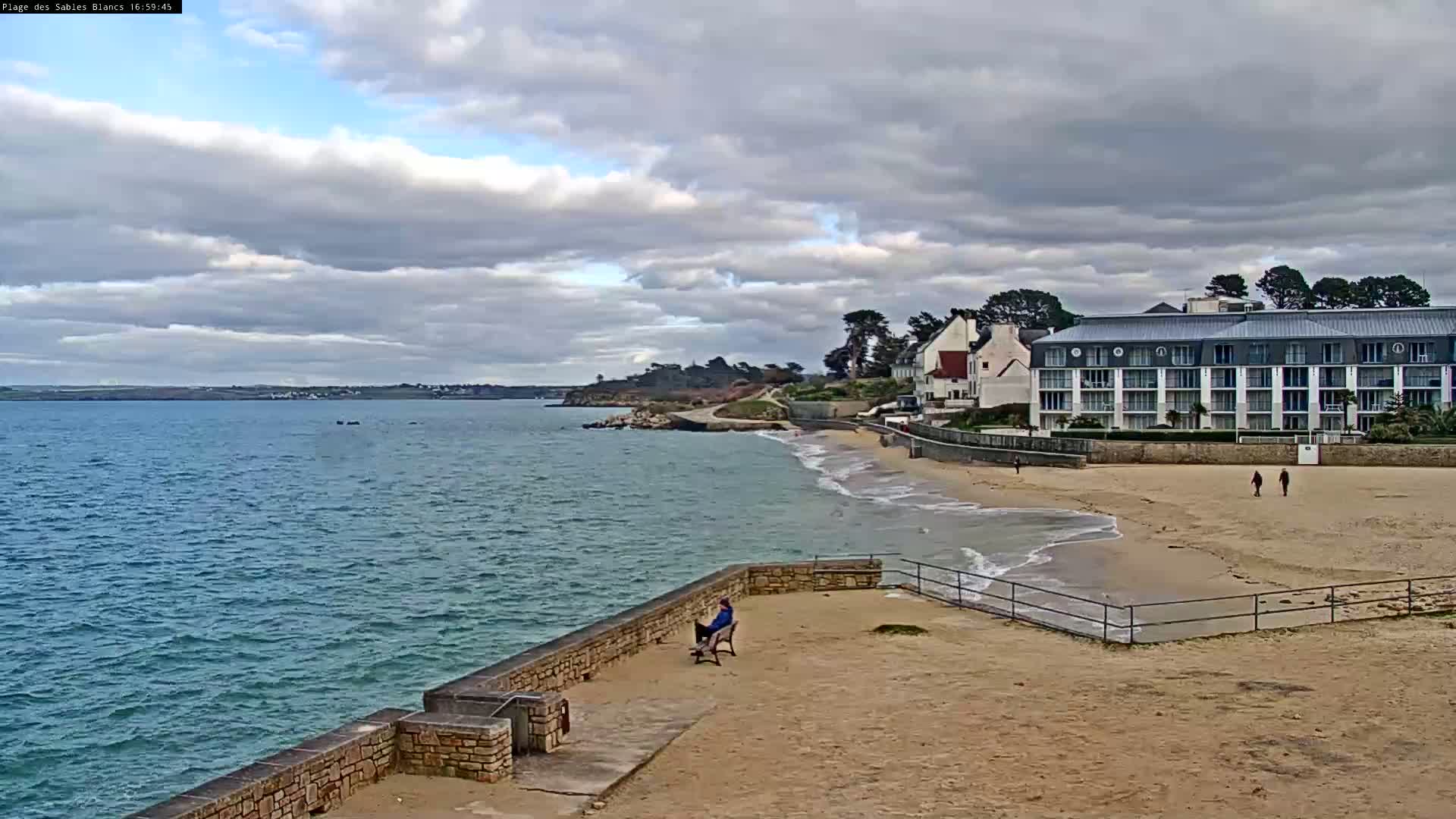 A person sits on a bench overlooking a sandy beach and calm ocean under a cloudy sky.