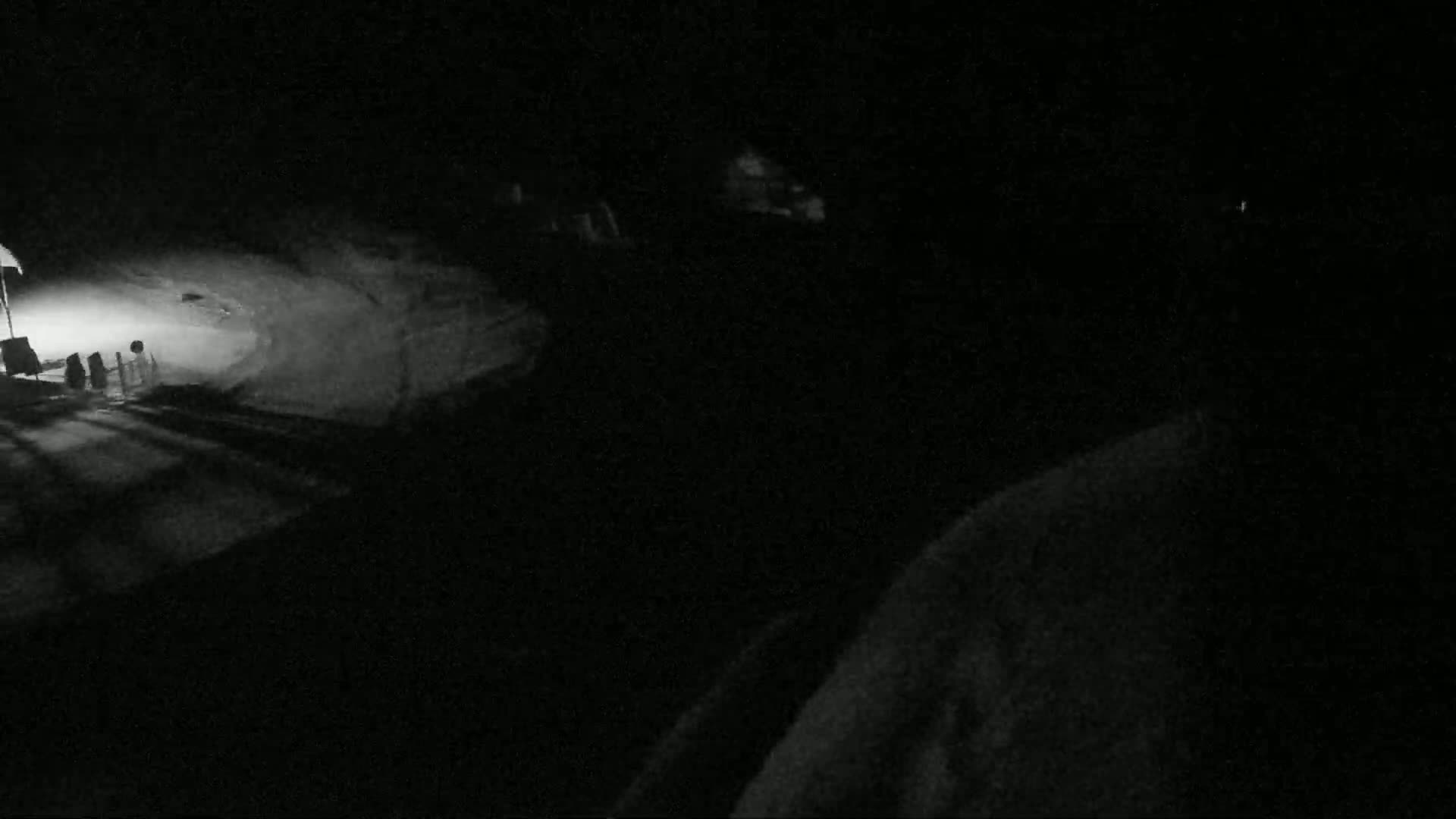 A dimly lit nighttime scene shows a snow-covered area with faint light sources and shadows cast on the ground.