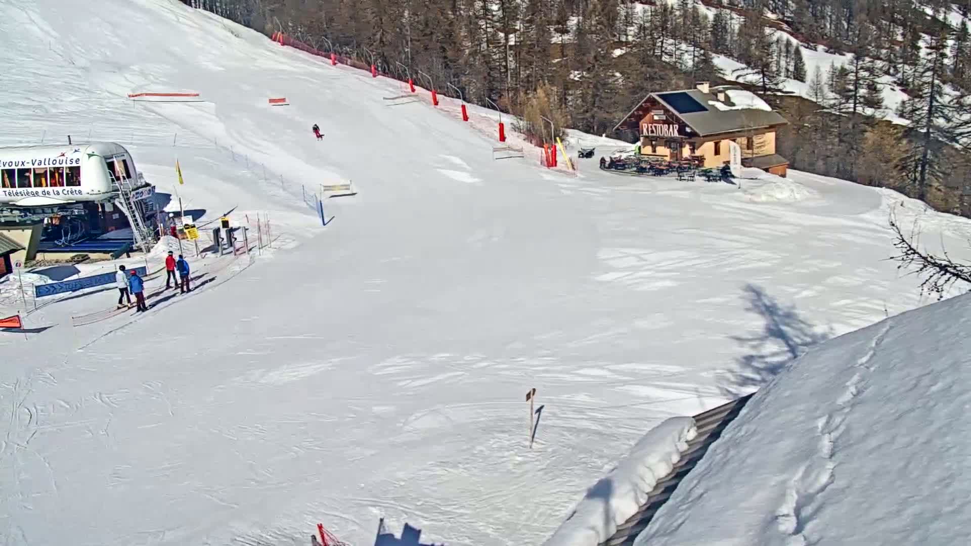 A snowy ski slope with a chairlift, a small building, and several skiers visible on a sunny day.