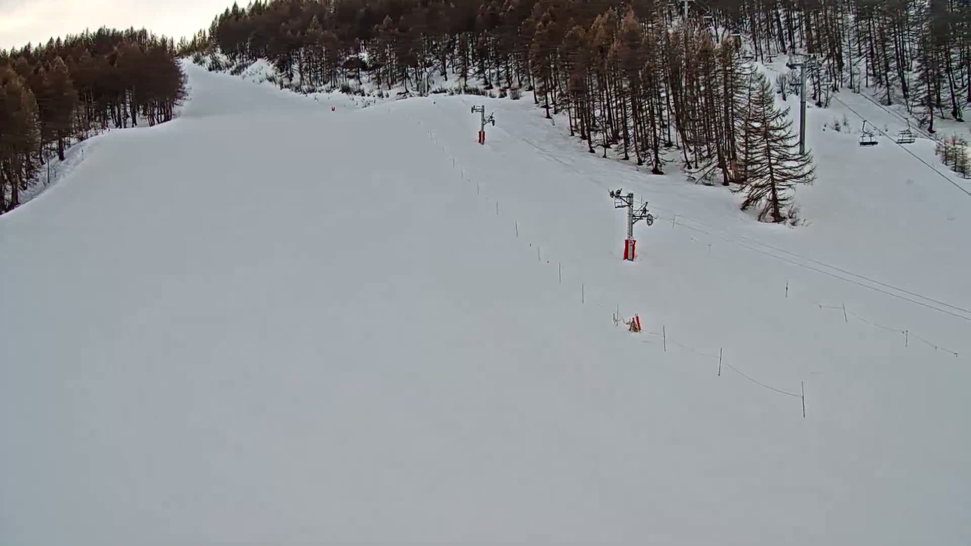 A snow-covered ski slope with two chairlifts is visible, bordered by a dark evergreen forest under a cloudy sky.