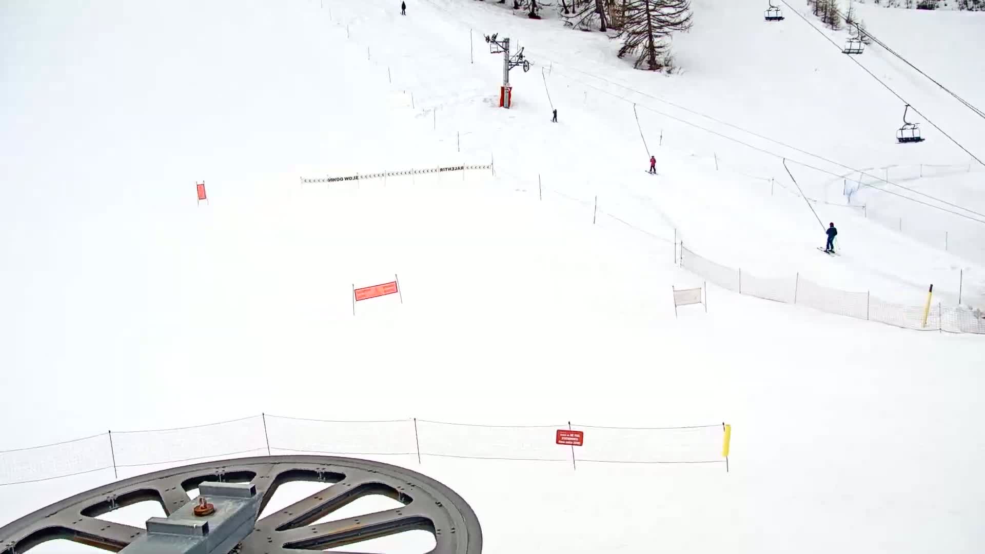 A large, metal pulley wheel is in the foreground, overlooking a snowy ski slope with several skiers and ski lifts.