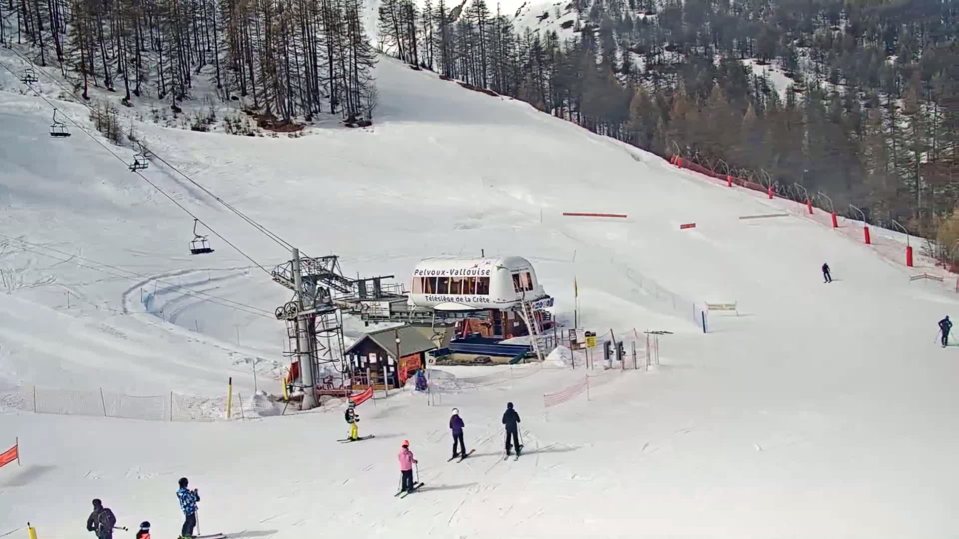 A ski lift and several skiers are visible on a snow-covered slope surrounded by trees under a bright, sunny sky.