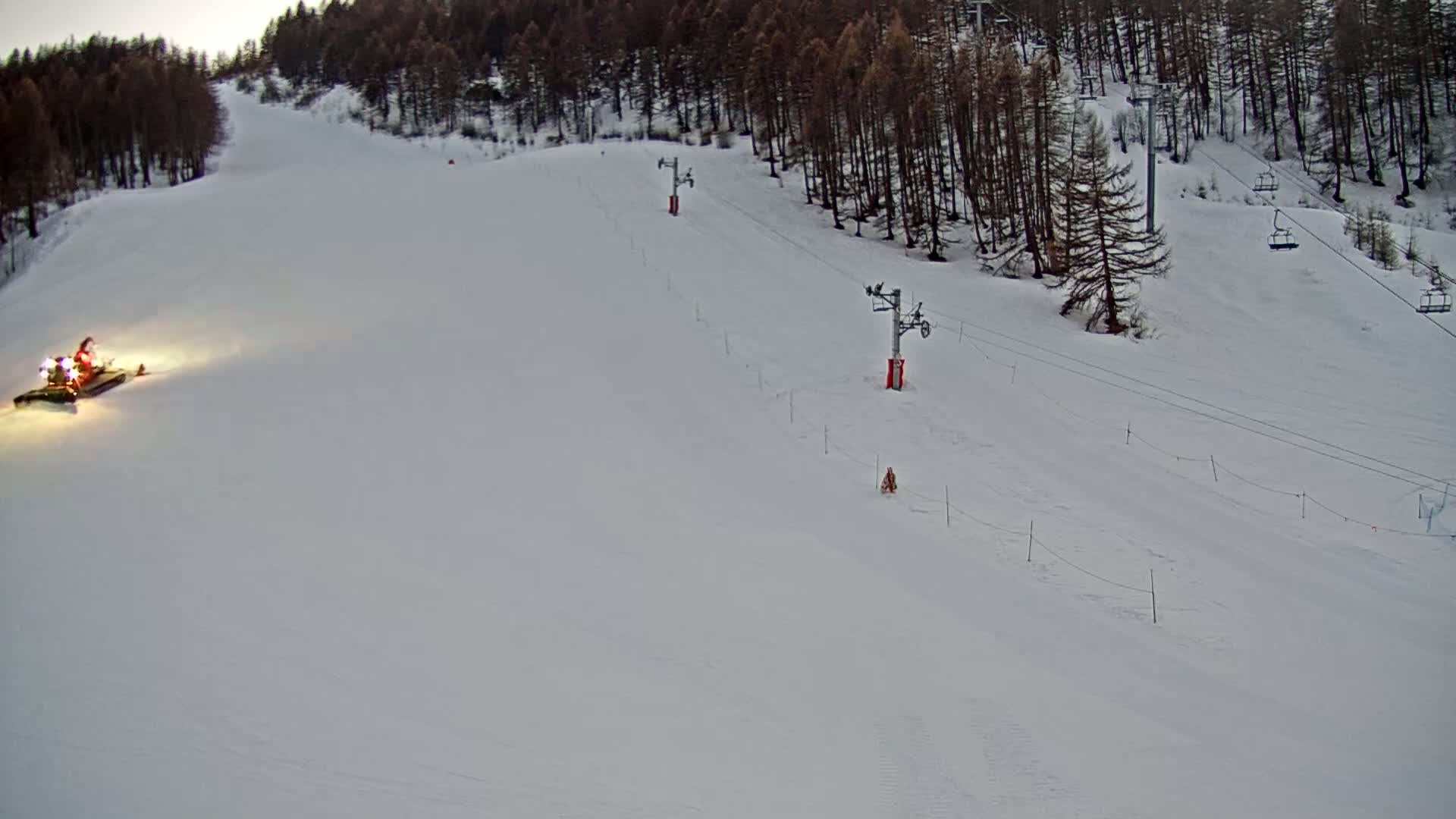 A snowcat grooms a snowy ski slope at dusk, with ski lifts and a pine forest visible in the background.