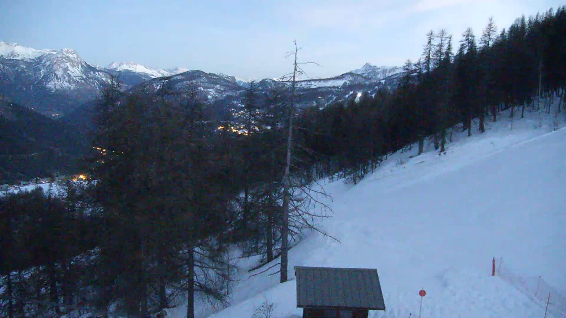 A snow-covered hillside with a small building and dark evergreen trees overlooks a valley with distant snow-capped mountains under a twilight sky.