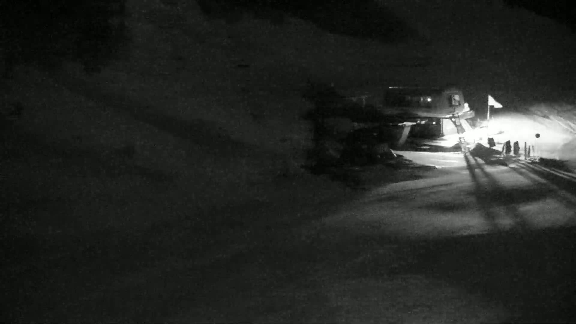 A snow-covered area at night is illuminated by headlights, revealing a building and several figures standing near a road.