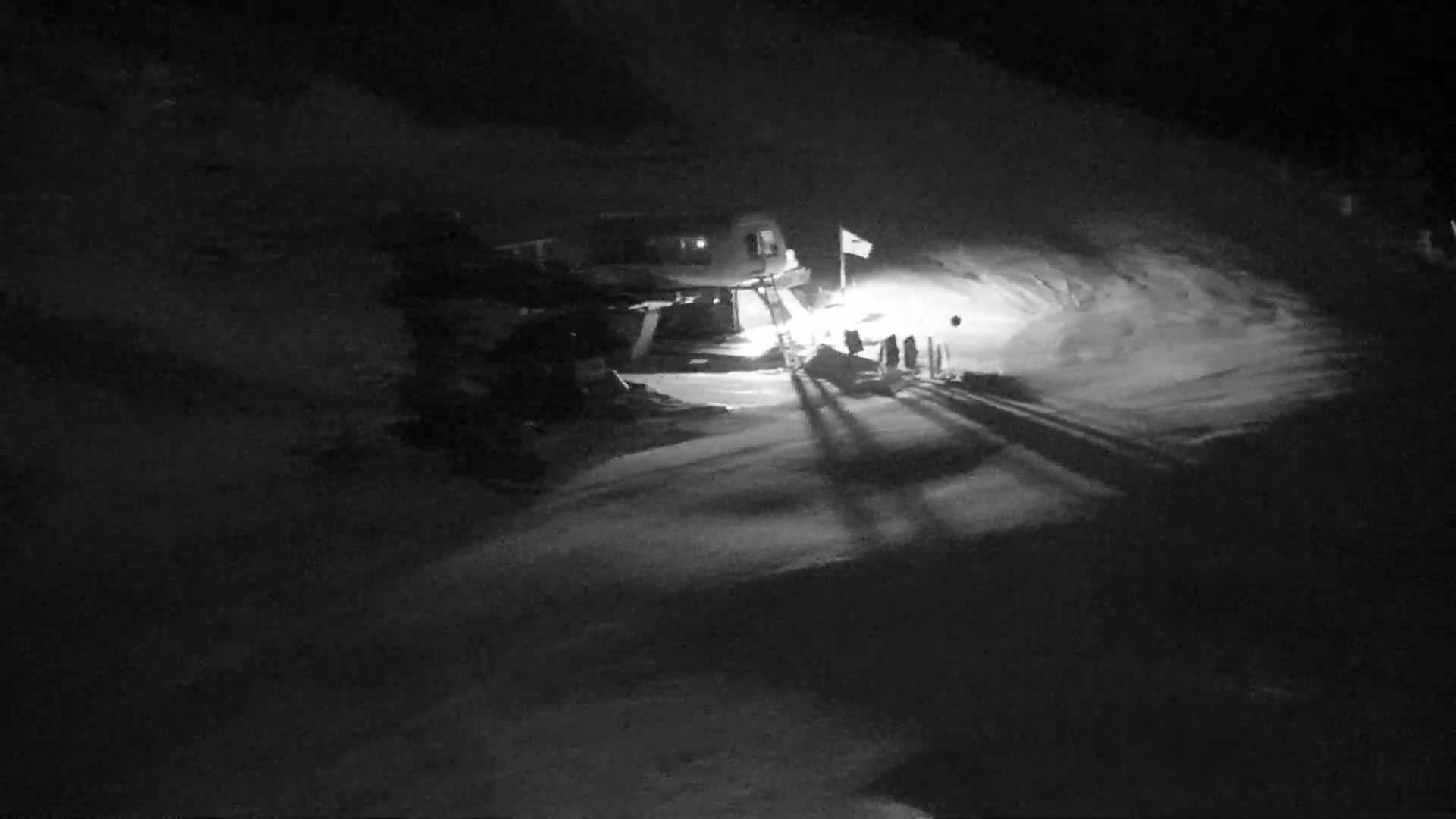 At night, a snow-covered area is illuminated, showing a building, a few people, and long shadows cast across the snow.