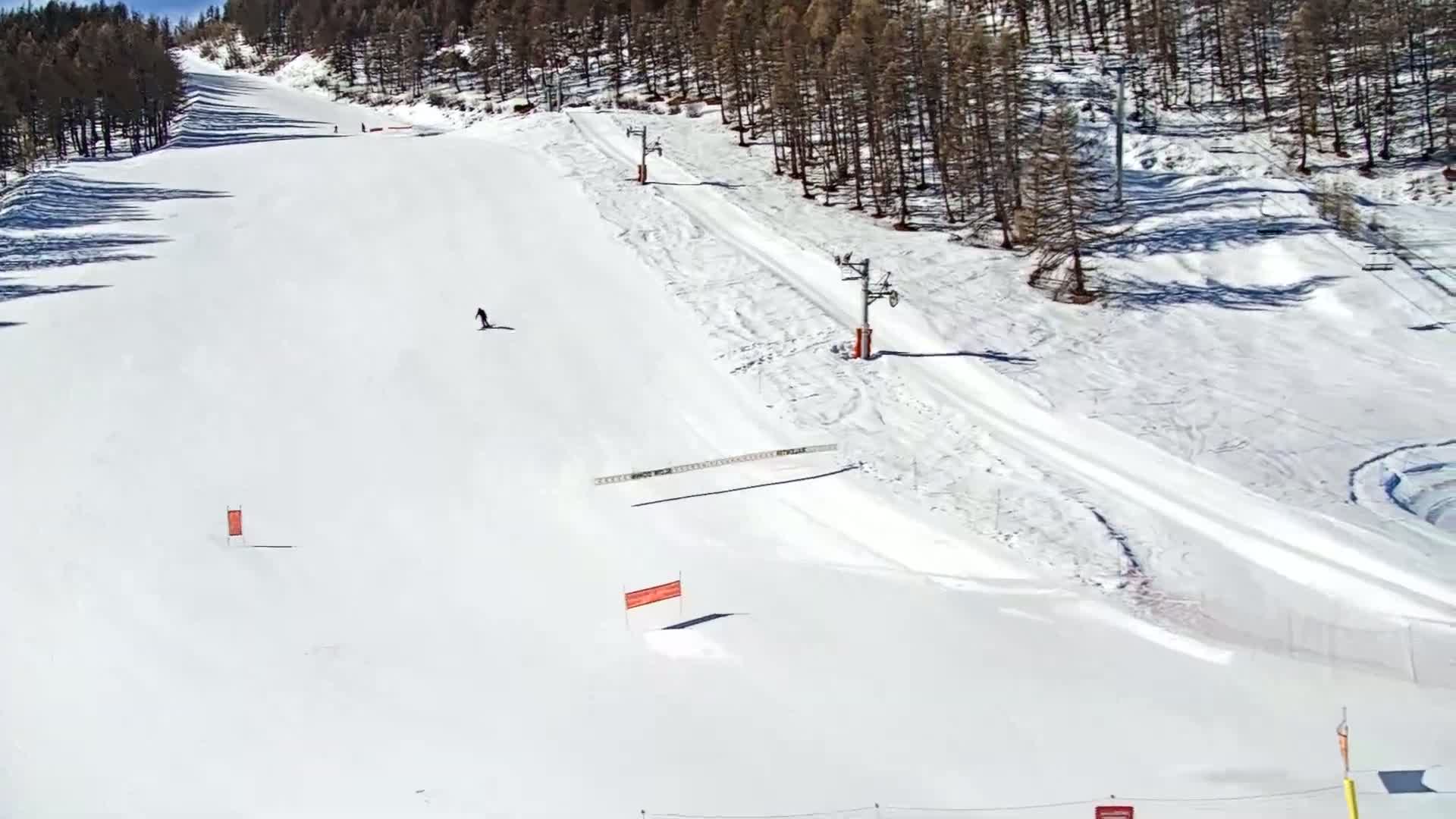 A snowy ski slope with two chairlifts, a few skiers, and orange markers is seen on a sunny day.