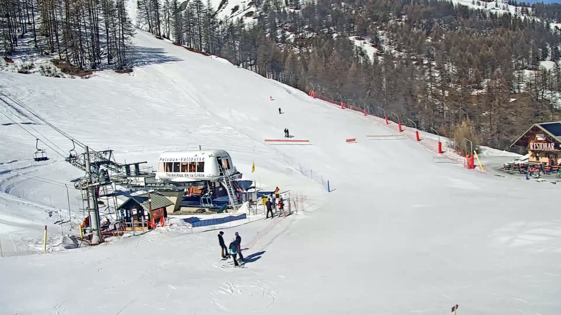 A ski lift and several skiers are on a snow-covered slope on a sunny day, with a forested hillside in the background.