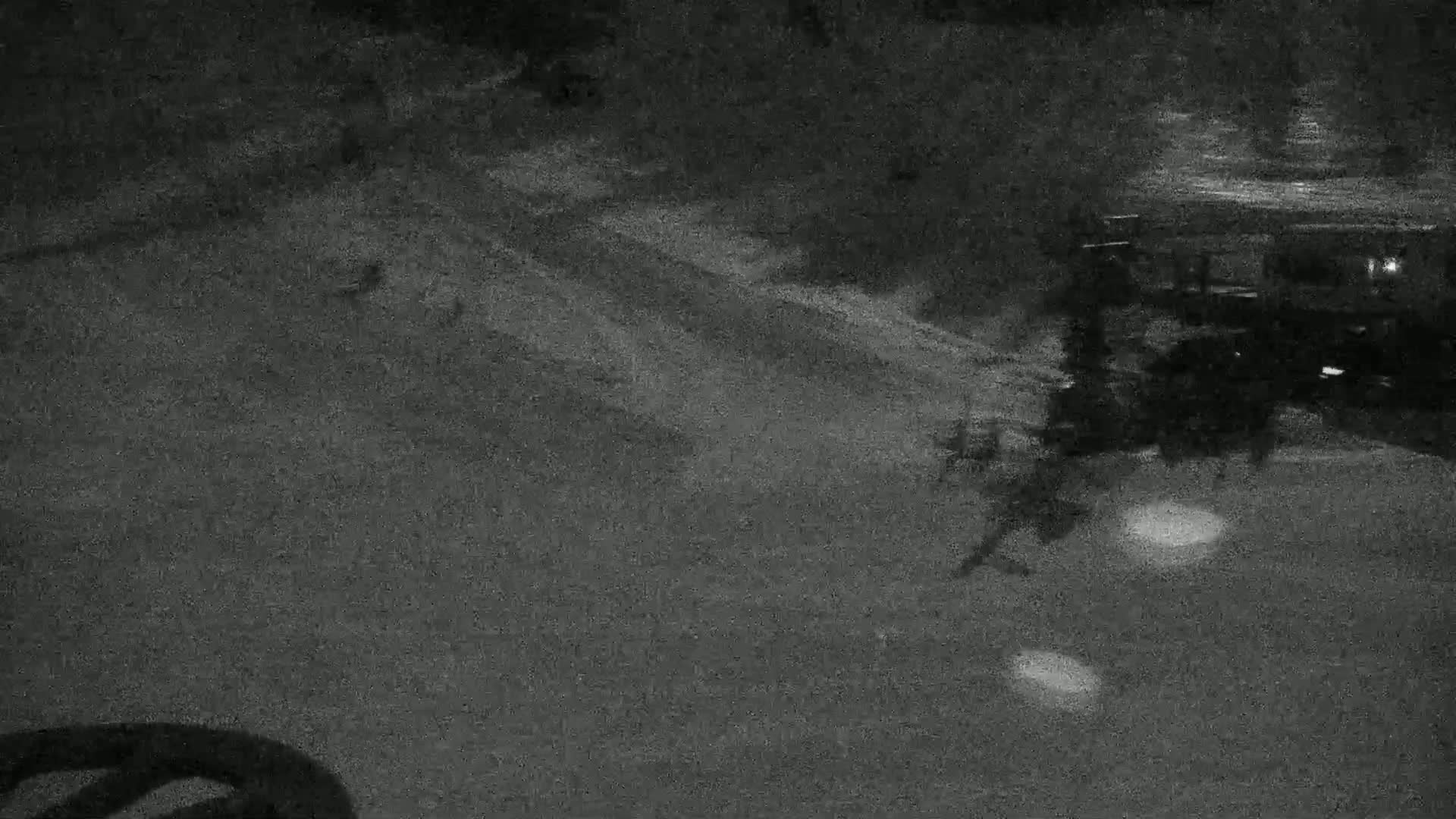 A nighttime, grainy image shows a dark, paved area with indistinct objects and shadows, possibly indicating outdoor furniture or vehicles, under low-light conditions.