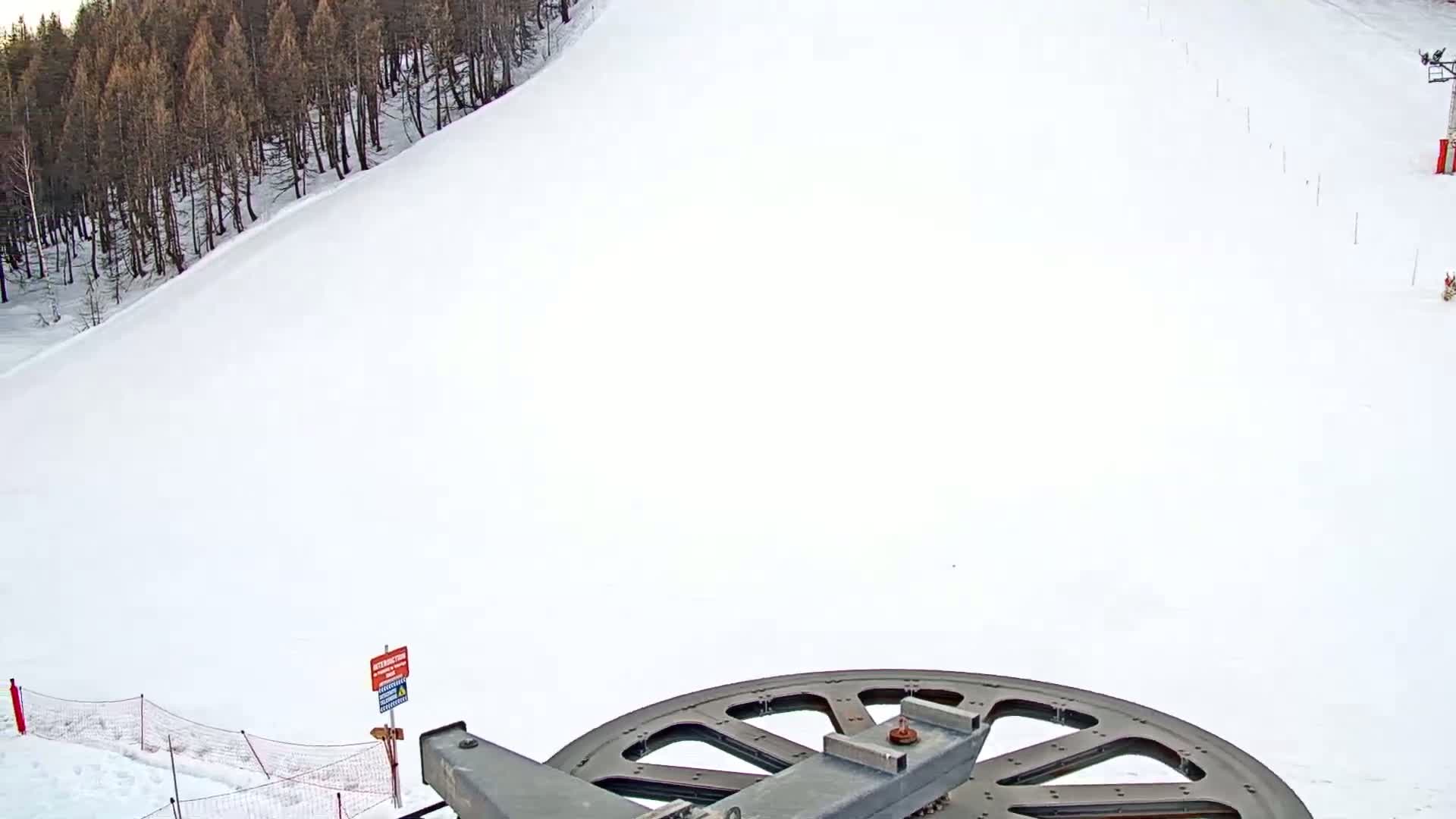 A snowy ski slope is seen from a low vantage point near a ski lift pulley, with a dark forest visible in the background.